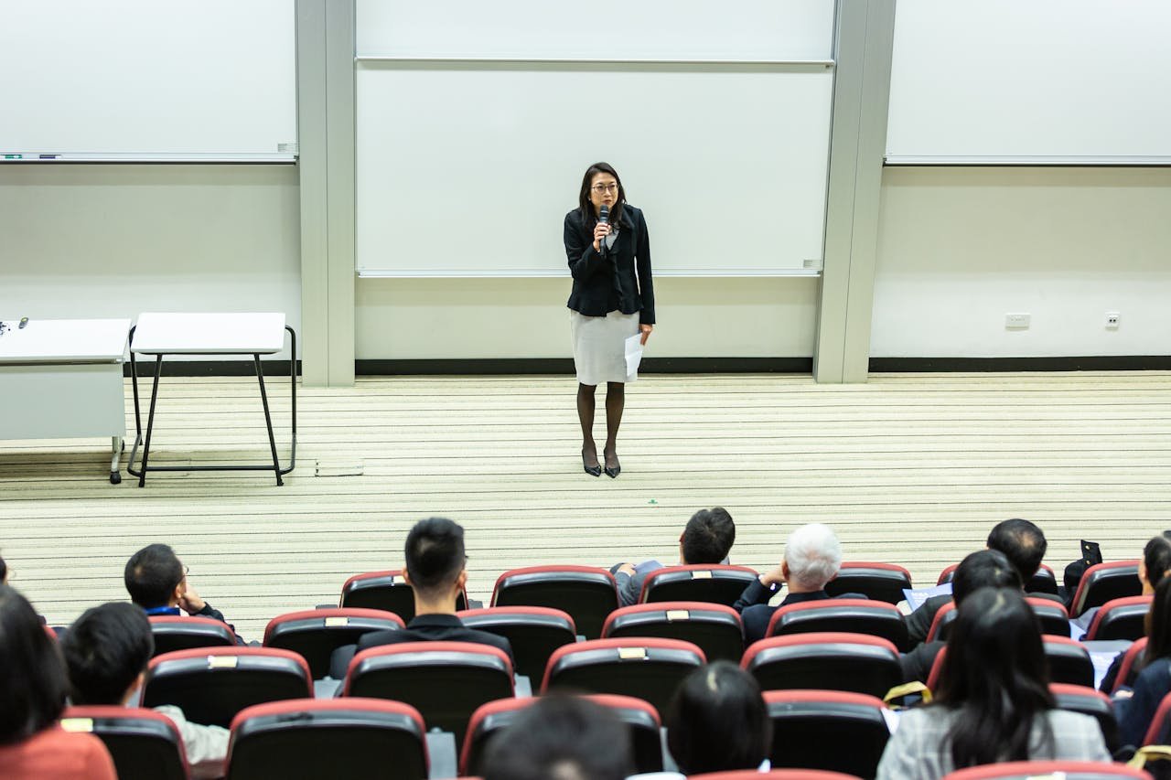 services-03 Female speaker presenting to an audience in a modern auditorium setting.